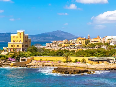Mediterranean seacoast in Alghero city, Sardinia, Italy. Spring flowers and trees on foreground, colourful buildings of Alghero old city center on background