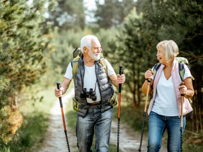 Happy senior couple hiking with trekking sticks and backpacks at the young pine forest. Enjoying nature, having a good time on their retirement